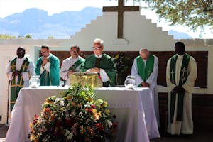 Elderly priest in eyeglasses and catholic vestment standing with prayer hands near table with Bible and multiethnic deacons during liturgy process behind mountains in sunlight