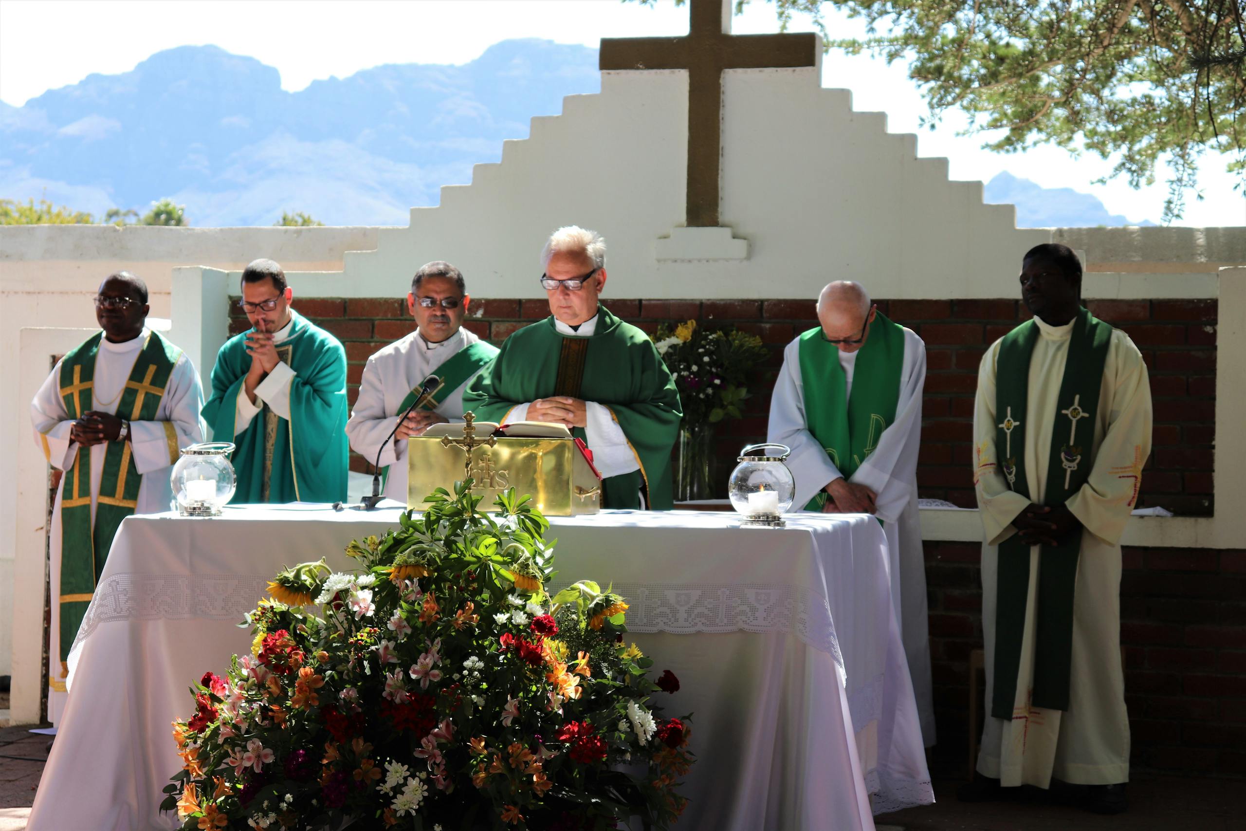 Elderly priest in eyeglasses and catholic vestment standing with prayer hands near table with Bible and multiethnic deacons during liturgy process behind mountains in sunlight